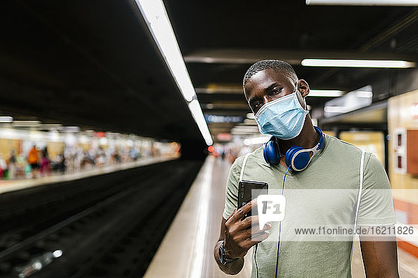 Junger Mann mit Maske benutzt sein Smartphone in einer U-Bahn-Station