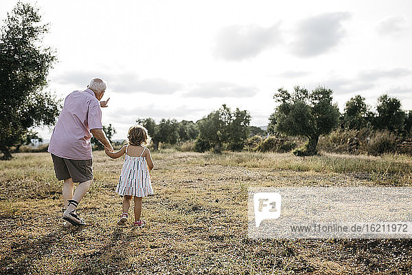 Grandfather with granddaughter walking on grassy land against sky