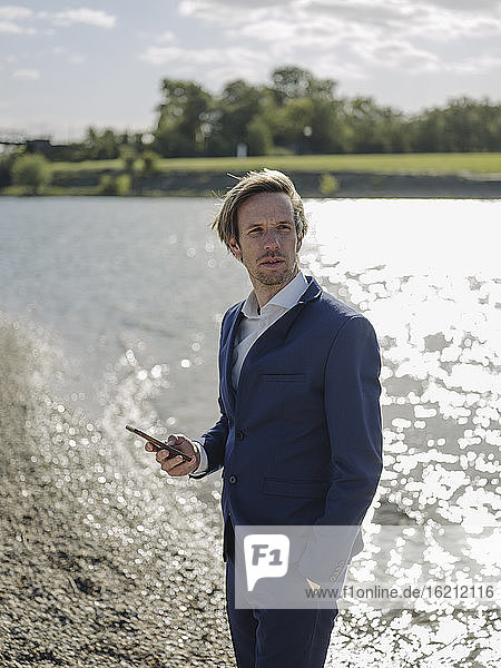 Businessman using smart phone looking away while standing at riverbank during sunny day