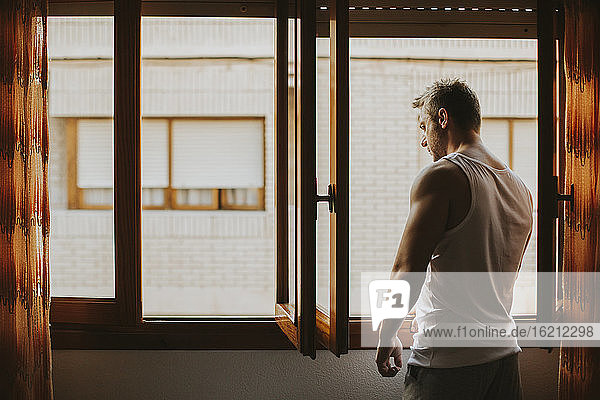 Thoughtful mid adult man looking through window while standing at home