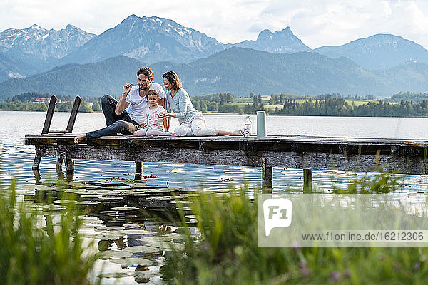 Eltern mit Tochter sitzen auf dem Steg über dem See vor den Bergen