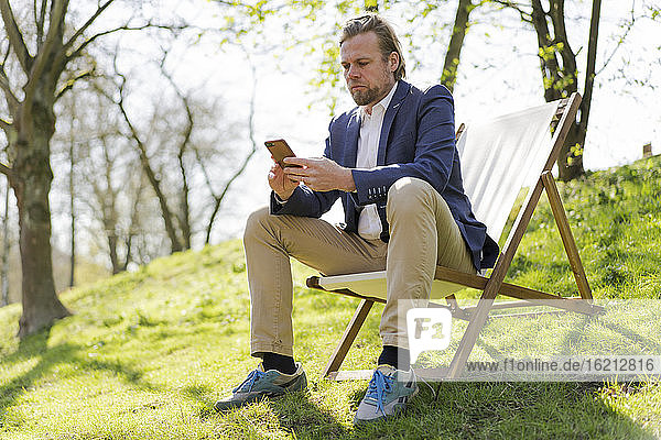 Männlicher Unternehmer  der sein Smartphone benutzt  während er auf einem Stuhl im Park sitzt