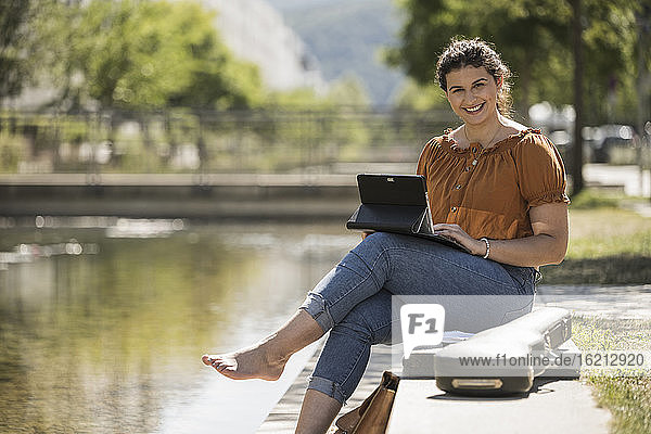 Lächelnde junge Frau  die einen Laptop benutzt  während sie an einem sonnigen Tag am Teich im Park sitzt