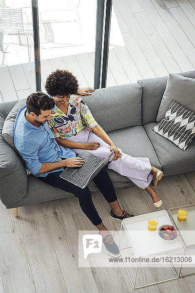 Multiethnisches Paar benutzt einen Laptop  während es auf dem Sofa im Penthouse sitzt