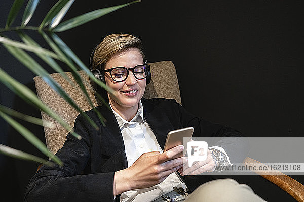 Lächelnde blonde schöne Geschäftsfrau  die ihr Smartphone benutzt  während sie sich entspannt und in der Pause im Büro Musik hört