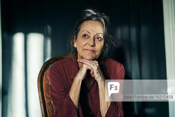 Nahaufnahme einer nachdenklichen älteren Frau  die zu Hause auf einem Stuhl vor einem Vorhang sitzt