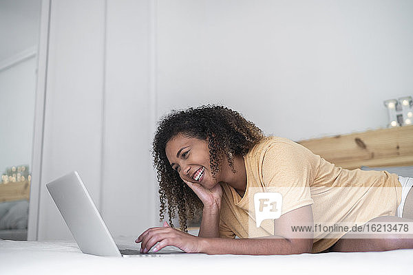 Fröhliche Frau mit lockigem Haar  die einen Laptop benutzt  während sie zu Hause auf dem Bett liegt