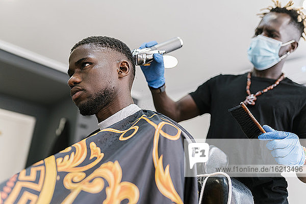 Junger Mann mit Maske und Handschuhen  der einem Kunden im Salon die Haare mit einem Rasiermesser schneidet