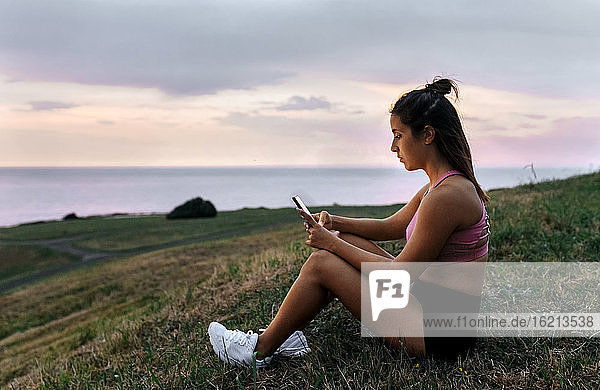 Junge Sportlerin benutzt ihr Smartphone,  während sie bei Sonnenuntergang im Gras sitzt
