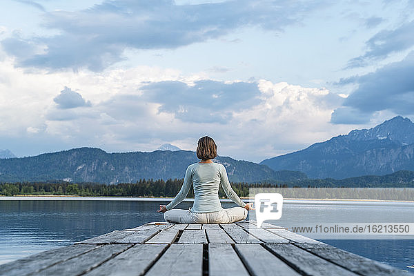 Mid adult woman meditating while sitting on jetty over lake against cloudy sky