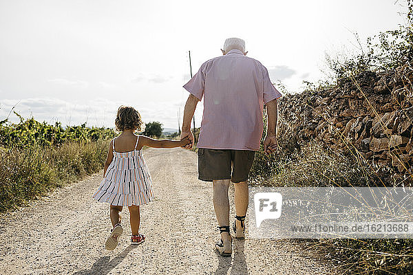 Grandfather holding granddaughter's hand while walking on dirt road against sky