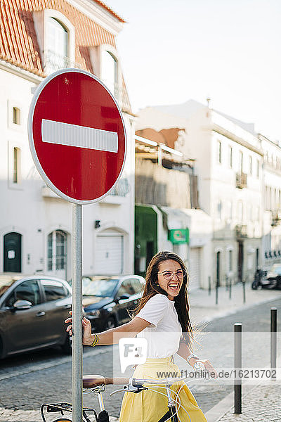 Fröhliche schöne Frau mit Fahrrad  die an einem Straßenschild in der Stadt steht