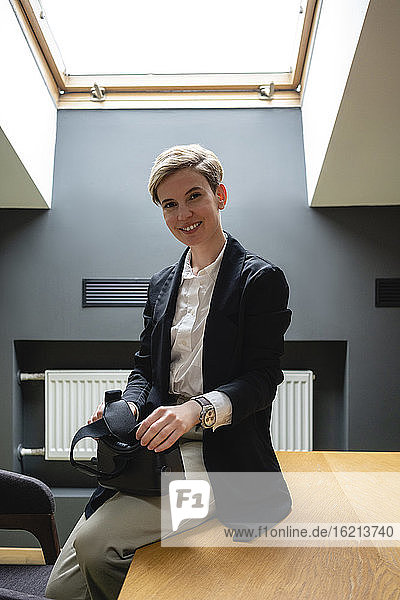 Lächelnde weibliche Fachkraft sitzt mit Virtual-Reality-Simulator-Headset auf dem Tisch im Büro