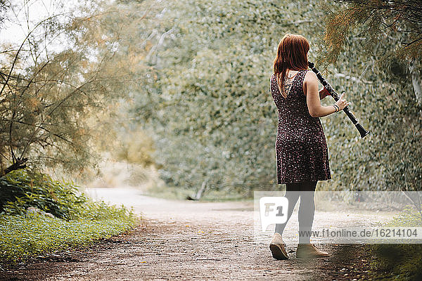 Junge rothaarige Frau  die Klarinette spielt  während sie auf einem Fußweg bei Pflanzen im Wald geht