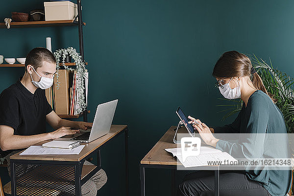 Junger Mann und Frau mit Schutzmasken bei der Arbeit an Schreibtischen zu Hause