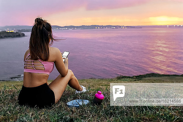 Sportlerin mit Smartphone im Gras sitzend mit Blick auf das Meer gegen den Himmel bei Sonnenuntergang