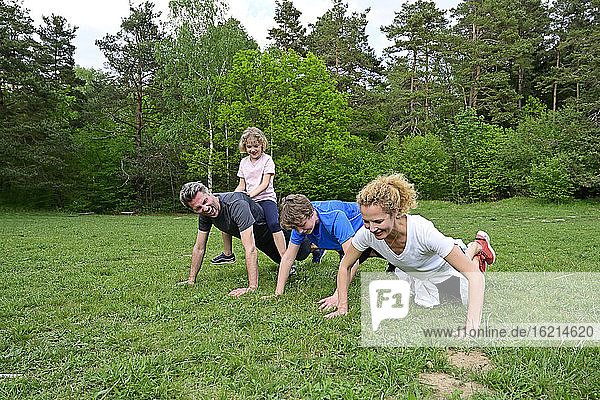 Girl sitting on father back doing push-ups with family over grassy land in forest