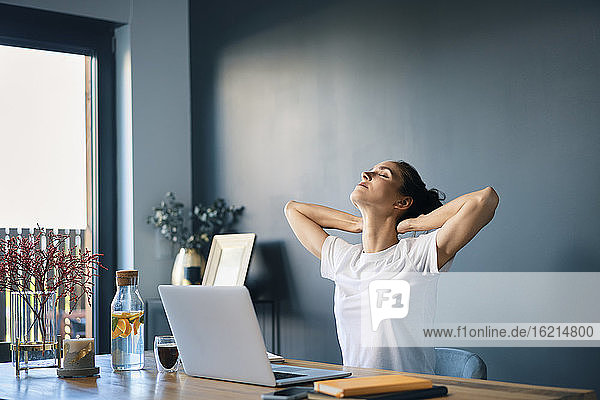 Müde Geschäftsfrau mit Händen hinter dem Kopf sitzt am Schreibtisch im Büro zu Hause