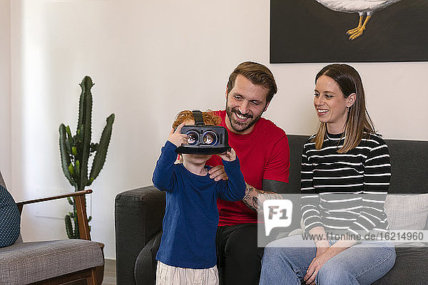 Lächelnde Eltern  die ihren Sohn in einem Virtual-Reality-Simulator im Wohnzimmer beobachten