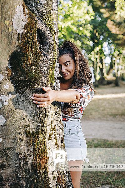 Smiling young woman holding tree trunk while standing in park