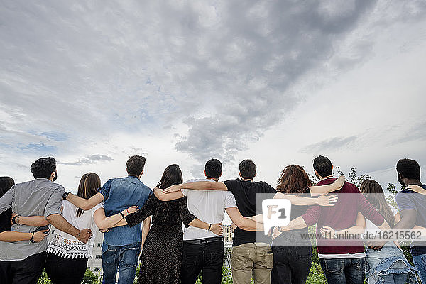 Rear view of female and male friends with arms around standing against cloudy sky