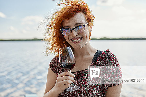 Porträt einer fröhlichen rothaarigen jungen Frau  die an einem See Wein trinkt