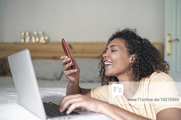Fröhliche junge Frau mit Smartphone und Laptop auf dem Bett zu Hause