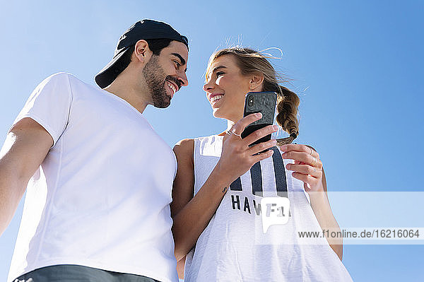 Lächelnde Frau  die ihr Smartphone hält und ihren Freund gegen den klaren blauen Himmel ansieht