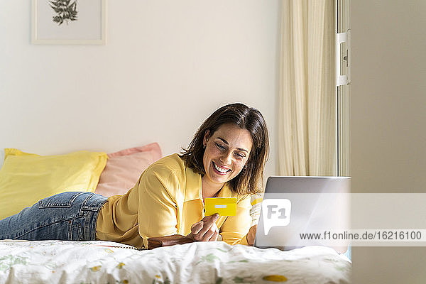 Lächelnde Frau schaut auf ihre Kreditkarte  während sie einen Laptop im Schlafzimmer benutzt