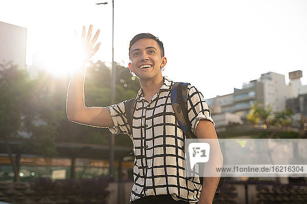 Smiling young gay man waving while standing in city against sky