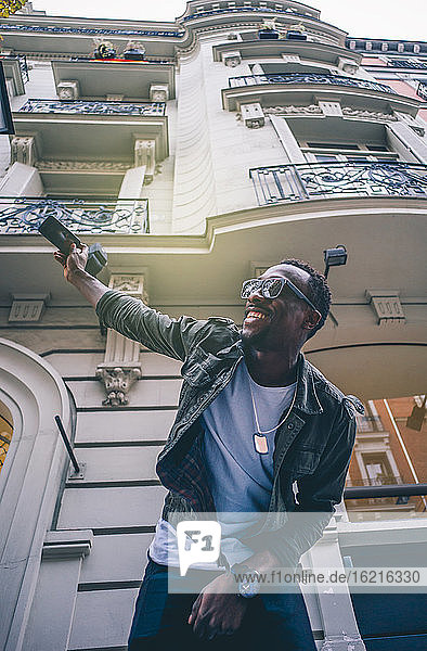 Smiling man wearing sunglasses taking selfie with smart phone against building in city