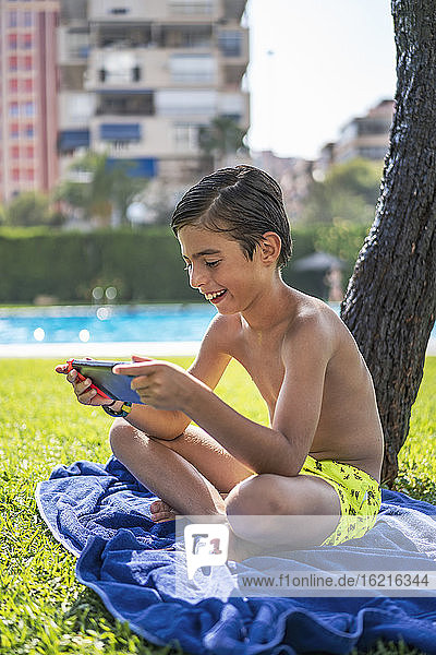 Hemdloser Junge spielt ein Videospiel auf einem digitalen Tablet  während er auf einem Handtuch im Hof sitzt