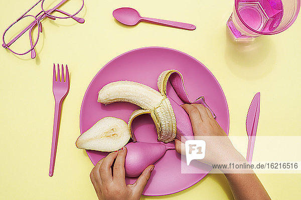 Hand of baby girl picking up pink-colored pear and banana from plastic plate