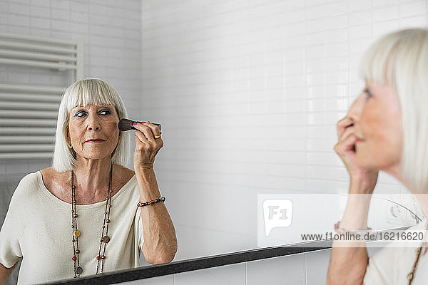 Ältere Frau trägt Make-up auf  während sie zu Hause in den Spiegel schaut