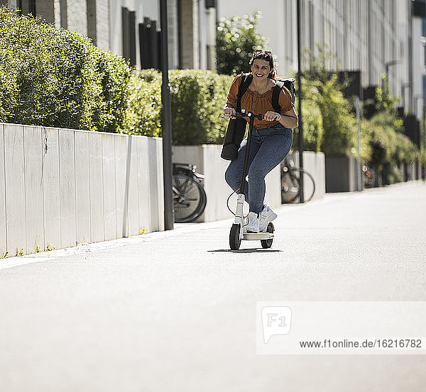 Lächelnde Frau auf elektrischem Roller auf der Straße an einem sonnigen Tag