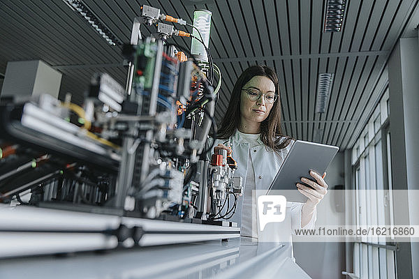 Junge Frau benutzt ein digitales Tablet  während sie an einer Maschine im Labor steht