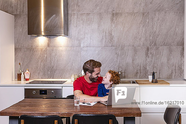Lächelnder Vater mit Sohn  der einen Laptop am Esstisch in der Küche benutzt