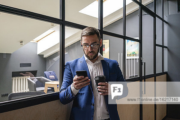 Businessman holding disposable coffee cup while using smart phone against glass wall at coworking office