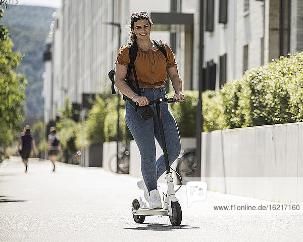 Lächelnde junge Frau auf elektrischem Roller auf der Straße an einem sonnigen Tag