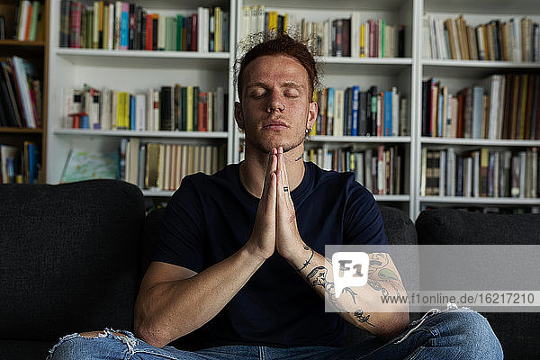 Man meditating while sitting against bookshelf at home