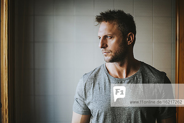 Thoughtful man looking through window while standing against wall at home