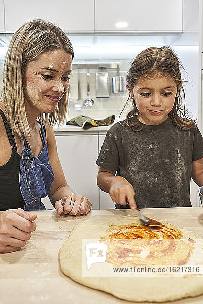 Mother looking at daughter preparing pizza on table in kitchen