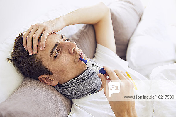 Close-up of sick man with thermometer in mouth resting on bed at home
