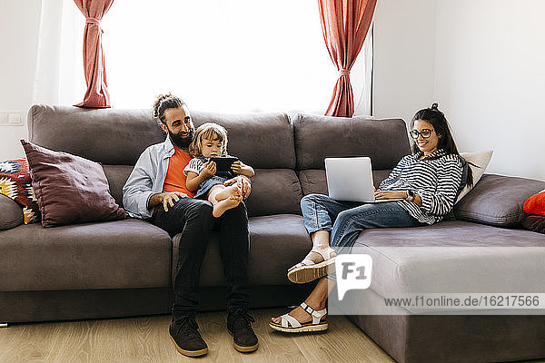 Frau arbeitet am Laptop  während Vater und Tochter auf dem Sofa im Wohnzimmer ein Mobiltelefon benutzen