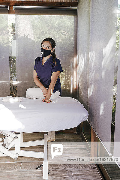 Weibliche Therapeutin mit Maske steht am Massagetisch im Spa