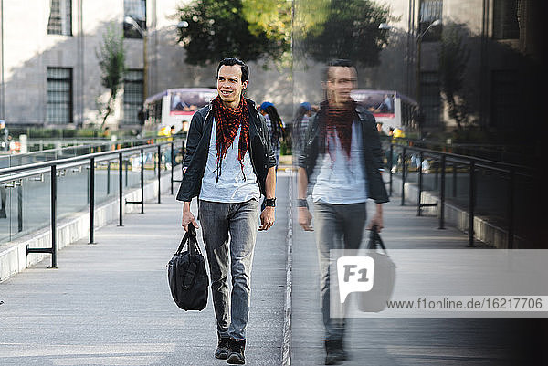 Selbstbewusster  modischer Mann  der sich auf einem Glas spiegelt  während er auf einem Fußweg in der Stadt spazieren geht
