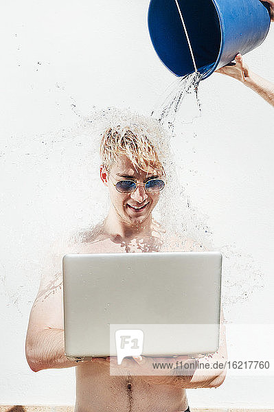 Hand einer Frau  die Wasser auf einen hemdlosen jungen Mann gießt  der einen Laptop an der Wand benutzt