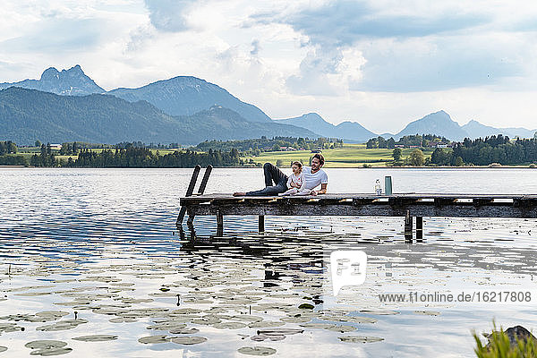 Vater und Tochter betrachten die Aussicht  während sie sich auf dem Steg über dem See vor den Bergen entspannen