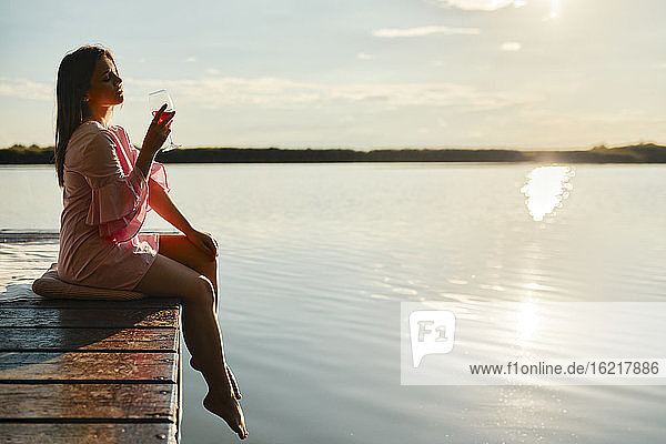 Junge Frau mit einem Glas Wein auf einem Steg an einem See bei Sonnenuntergang sitzend