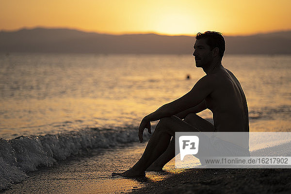 Nachdenklicher Mann ohne Hemd  der bei Sonnenuntergang am Ufer sitzt und wegschaut